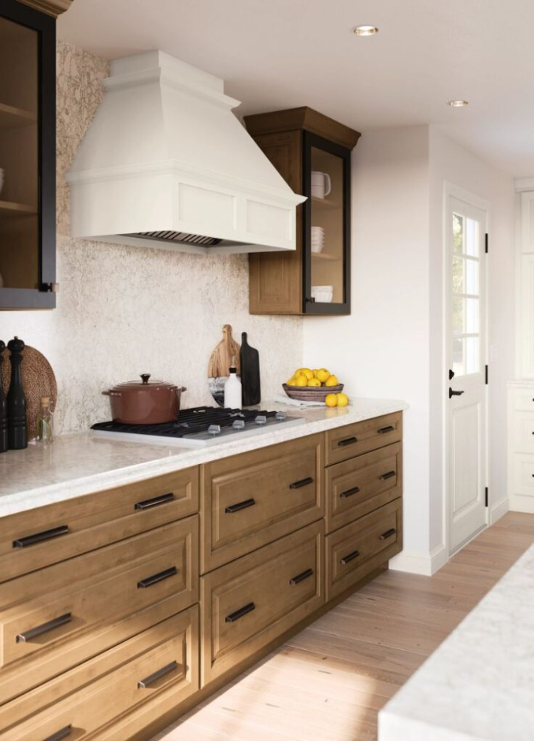 Farmhouse kitchen with wood and white cabinets, cream range hood, gas stovetop, cutting boards, lemons, and potted plants.