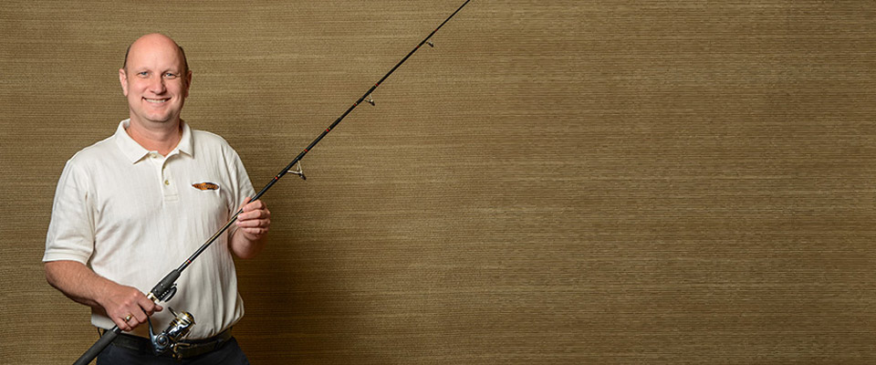 Don Hannah wearing a white polo shirt, standing in front of a gold backdrop and holding a fishing pole.